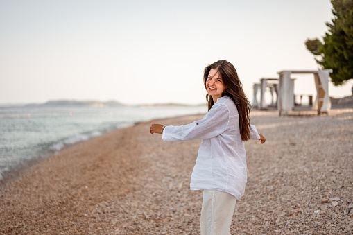 Young teenage girl with long dark hair, wearing white lined shirt enjoying a fresh morning on the empty beach in a summertime.