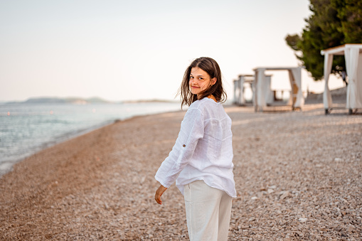 Young teenage girl with long dark hair, wearing white lined shirt enjoying a fresh morning on the empty beach in a summertime.
