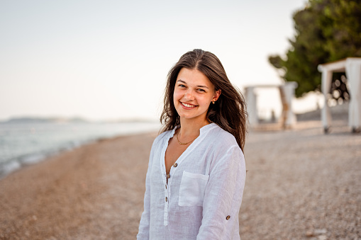 Young teenage girl with long dark hair, wearing white lined shirt enjoying a fresh morning on the empty beach in a summertime.