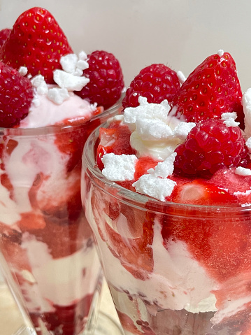 Stock image showing two knickerbocker glasses filled with Eton mess dessert made with layered pureed strawberries, whipped cream and crumbled meringue garnished with raspberries.