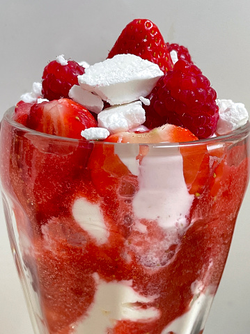 Stock image showing knickerbocker glass filled with Eton mess dessert made with layered pureed strawberries, whipped cream and crumbled meringue garnished with raspberries.