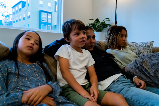 A living room portrait captures a diverse family's harmony on a comfortable sectional, the movie's allure uniting them. The youngest boy's brown hair and green shorts match his lively spirit. Next, a confident African American teenage girl with curly hair and blonde streaks stands out, flanked by two sisters in navy hoodie and blue floral dress, each a testament to their unique joy.