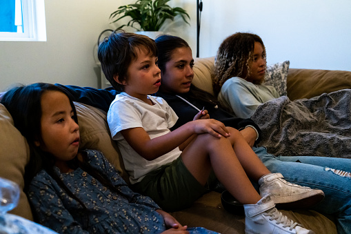 In a warm and welcoming living room, a diverse family has gathered on a spacious, plush sectional couch. The youngest boy, exuding joyful mischief, sports green shorts and sneakers, his gaze fixed on the screen.