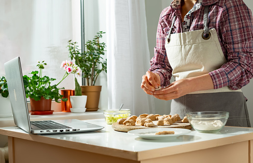 A woman prepares a dish from dough in the kitchen using a laptop, watching a video recipe on the computer.