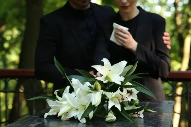 Couple near granite tombstone with white lilies at cemetery outdoors, selective focus. Funeral ceremony Couple near granite tombstone with white lilies at cemetery outdoors, selective focus. Funeral ceremony