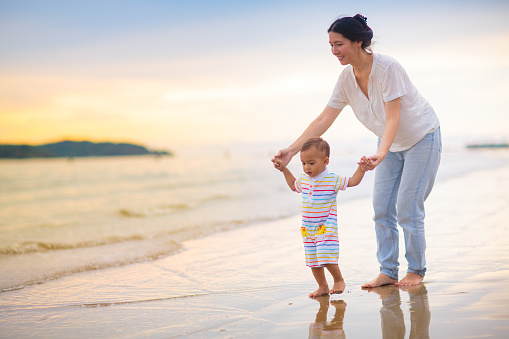 Mother and baby on tropical beach at sunset. Asian woman with infant boy walking and playing at sea. Young mom and little boy walk on ocean coast. Travel with kids. Vacation with young child.