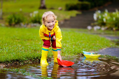 Child playing with paper boat in puddle. Kids play outdoor by autumn rain. Fall rainy weather outdoors activity for young children. Kid jumping in muddy puddles. Waterproof jacket and boots for baby.