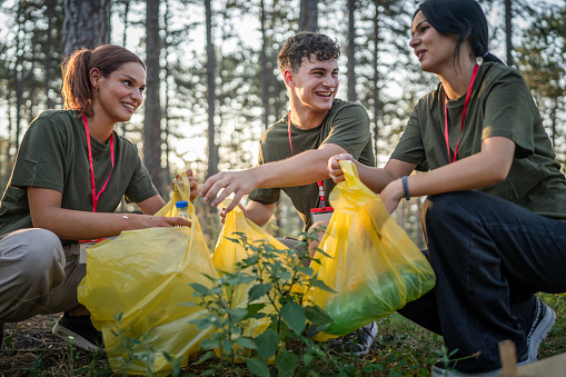 group of teenage friends gen Z male and female caucasian men women picking up waste garbage plastic bottles and paper from the forest cleaning up nature in sunny day environmental care ecology concept