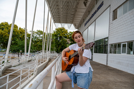 Side view of young female musician playing song on acoustic guitar while standing near metal fence of building on street in public place