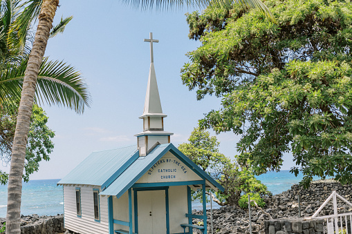 A small unique catholic church sits under palm trees on the coast of Kona. The ocean is in the background.
