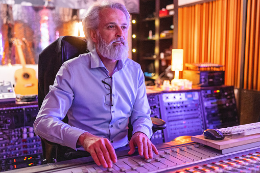 Portrait of a senior caucasian male sound engineer working on sound adjustiments while sitting in a recording studio. He is surrounded by sound recording and audio equipment. There is a guitar in the back and beautiful orange and purple lights that are illuminating the room. The sound engineer is adjusting the sound on the music mixer and looks serious. He has grey hair and a beard.