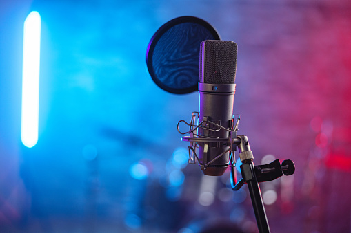A close up of a microphone on a music stand in a cool recording studio. The recording studio is illuminated by beautiful red and blue lights. There is a pop filter on the microphone while drums are partially visible in the background.