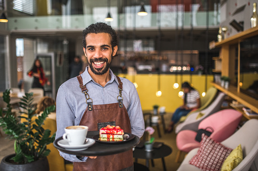 Portrait of a cheerful Middle Easter waiter standing in a cafe holding a serving tray with food and drinks on it.