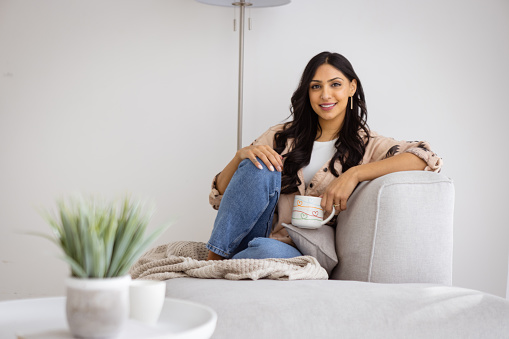 Beautiful Indian Woman Relaxing at Home With a Large Cup in her hands, sitting on a sofa in a sunny living room