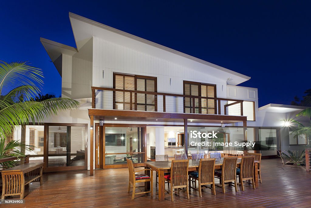Contemporary two story home with deck at dusk A luxurious house at dusk with a wooden entertaining area.  The exterior of the two-story house has many windows and open spaces.  A dining table with chairs on a large wooden deck is in the foreground.  Palm trees are on the edges of the deck.  The house is white with a slanted roof. House Stock Photo