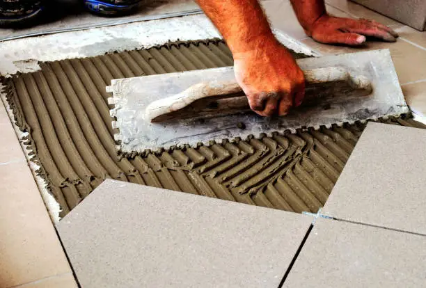 man's hands spreading cement mortar grout on concrete base for tiling. metal trowel tool. laying down large ceramic floor tiles. work in progress. construction work process.