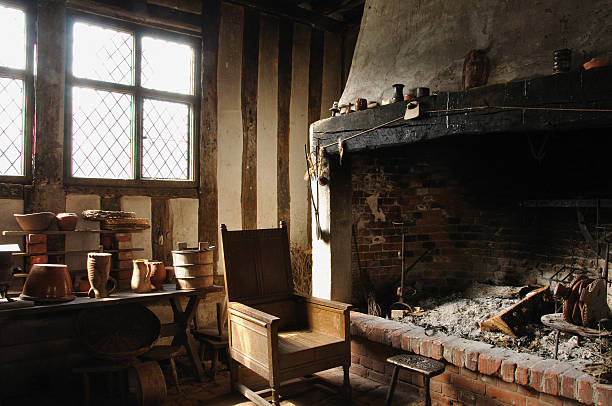 An empty wooden chair sits next to a large open fireplace full of ash.  The tudor beamed room with light streaming through the leaded windows creates a romatic and quiet mood which typifies a much older, rustic and basic way of life.  Suffolk, England.