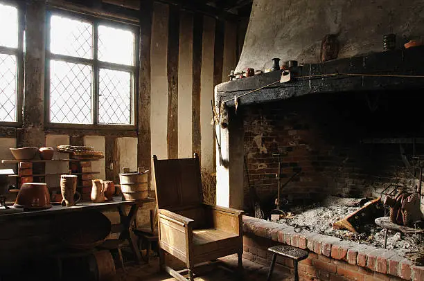 An empty wooden chair sits next to a large open fireplace full of ash.  The tudor beamed room with light streaming through the leaded windows creates a romatic and quiet mood which typifies a much older, rustic and basic way of life.  Suffolk, England.