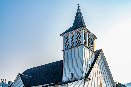 horizontal image of a quaint little white country church with a steeple sitting in a green meadow surrounded by trees under a beautiful blue sky with white clouds floating by in the summer time.