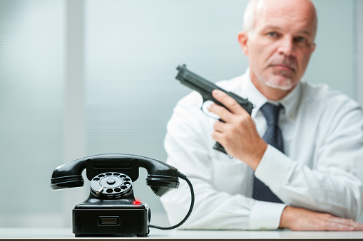 Phone clearly visible, hitman with pistol blurred. Represents a professional killer in a business office, suggesting a deep-rooted system link. Dressed as a senior executive