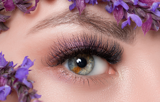 Woman's blue green eye with pupil heterochromia with beautiful make-up. Near her are beautiful blooming spring flowers. Looking at the camera. Professional makeup and cosmetology.