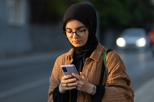 Pretty hijab lady is typing a message while walking on the street.