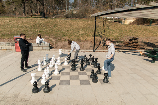 Two teenage boys are playing chess match against one man in city park. They are playing street cheese with huge table board and figurines.