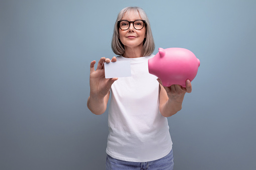 savings account. mature woman with gray hair keeps her savings on a credit card and in a piggy bank on a studio background with copy space.