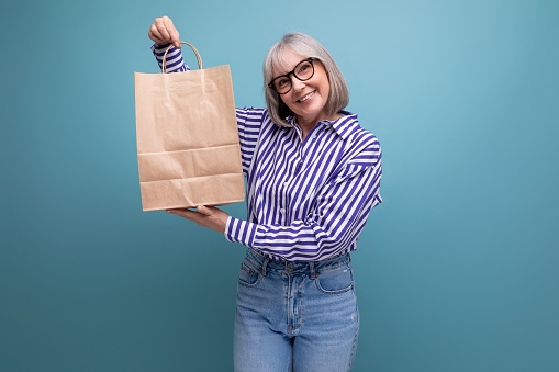 fashionable middle-aged woman with gray hair holding out a package with social assistance on a bright studio background with copy space.