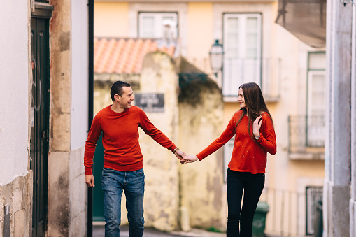 man and woman in stylish red sweaters hold hands and walk. city street with beautiful buildings.