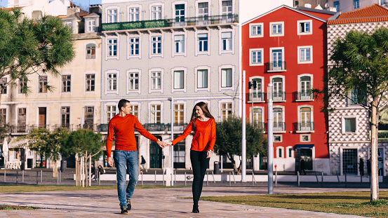 guy and girl in stylish red sweaters hold hands and look at each other. large square in front of buildings.