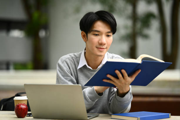istock Peaceful young asian man student reading interesting book while sitting bench in sunny summer park. 1639816643