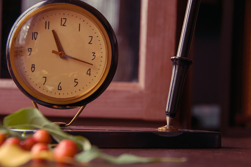 Vintage desk clock with ink fountain pen and apple tree leaves with small apples on wooden background with bokeh. Autumn season picture style.
