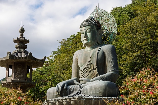 A large statue of Buddha seated atop a cement rock monument in front of a lush green mountain in Korea