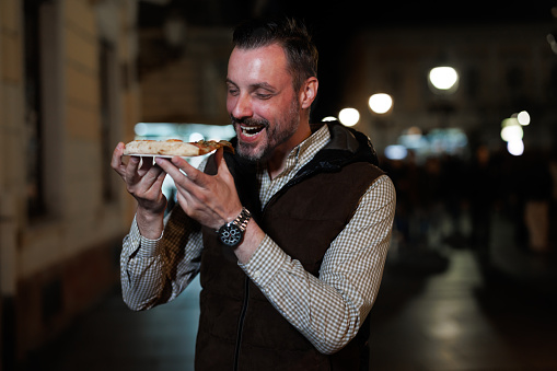 Under the captivating night sky, a man indulges in a moment of pure culinary pleasure, relishing the deliciousness of a slice of pizza on the bustling city street, savoring each bite with contentment