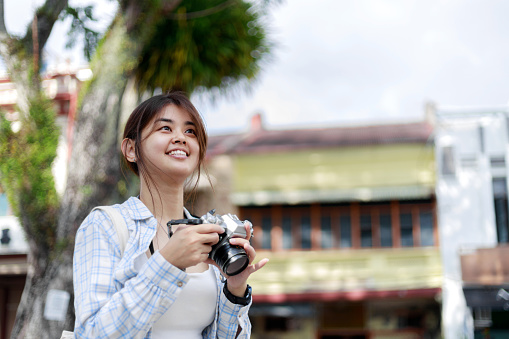 The happy Asian young woman is exploring and walking through a local city street while using her vintage film camera to document her surroundings. She's likely enjoying the sights and capturing moments as she explores the city.