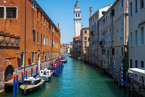 Venice, Italy – June 16, 2023: Beautiful summer day with colorful boats docked along the canal with a bell tower in the background in Venice, Italy.