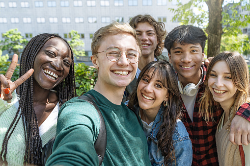 A group of multicultural teenagers with backpacks take a selfie in their school park. One has headphones around his neck. They're all smiling.
