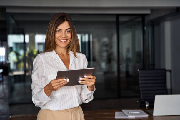 Latin Hispanic 40s years mature business woman looking at camera. smiling European businesswoman CEO holding digital tablet using fintech tab online application standing in modern office, copy space stock photo