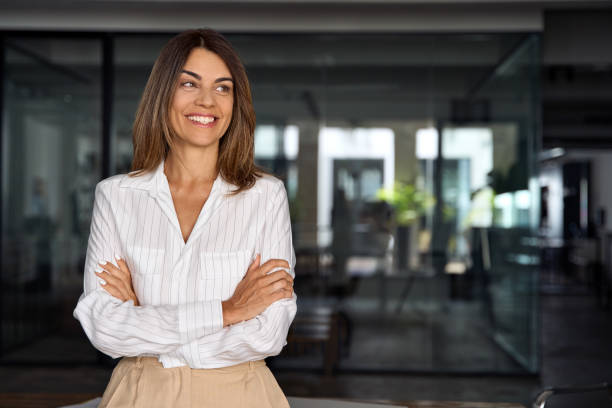Beautiful hispanic senior business woman with crossed arms smiling, looking aside. European Latin confident mature good looking middle age leader female businesswoman on office background, copy space. stock photo