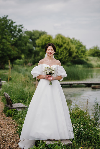 Portrait of bride holding a bouquet white flowers roses and gypsophila in nature. Young beautiful bride in a dress on shore of a lake outdoors. Happy wedding day of marriage. Getting married outdoors.