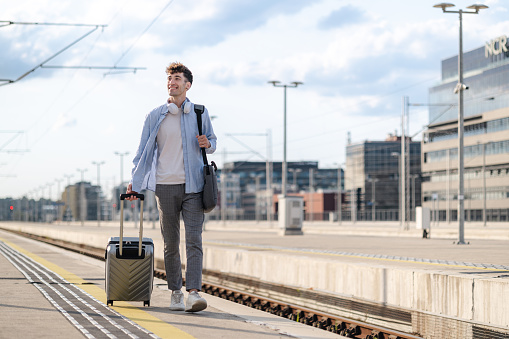 Full length of a young man walking on a train station with a carry on suitcase