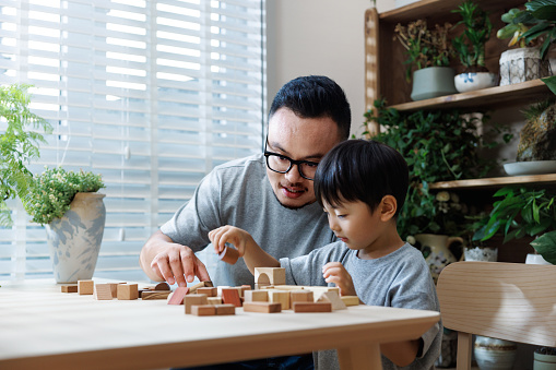 This is an Asian father and son, the father is about 35 years old and the son is about 3 years old. They are wearing casual clothes and sitting on the balcony at home, playing with building block toys together.