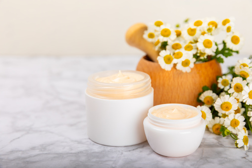 Open jar of moisturizing cream for face, body and hands with a chamomile flower on a light background. Herbal dermatological cosmetic hygiene cream. Natural cosmetic product. Beauty concept. MOCKUP