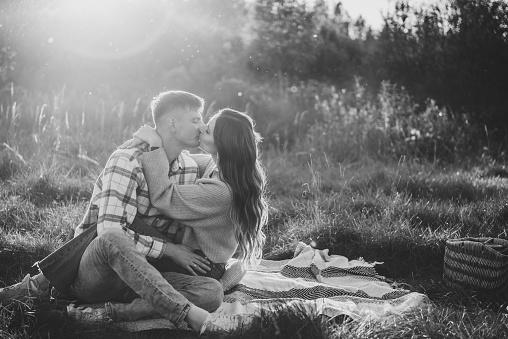 Couple sitting on blanket hugging in grass in field at sunset. Happy young woman kiss man spending time together in nature. Concept family holiday. Female embrace male on picnic. Black and white photo