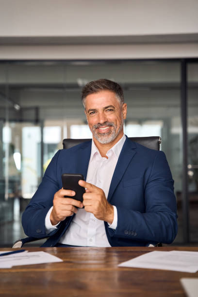 istock Vertical portrait of middle age Hispanic business manager using cell phone mobile app. Smiling Latin mature man businessman looking at camera, holding smartphone sit in office working online on gadget 1640251972