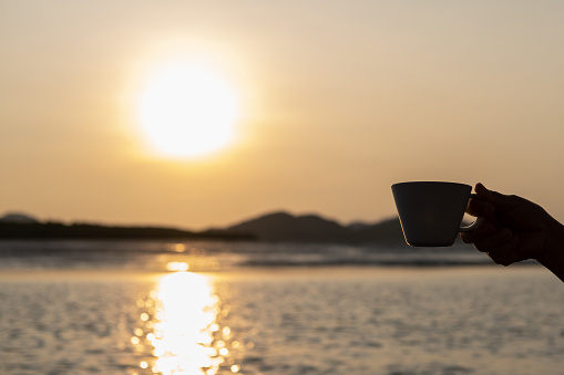 Hand holding a cup of coffee, with sunrise and sea view in the background