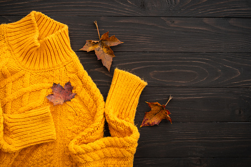 Cozy autumn background. Knitted sweater with fall leaves on wooden rustic background. Flat lay image.