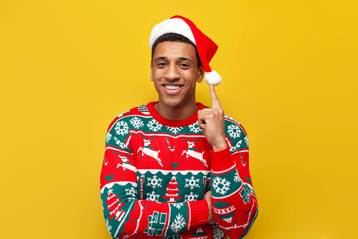 african american guy in New Year's clothes and santa hat smiles on yellow isolated background, man raises his hand up and points his finger