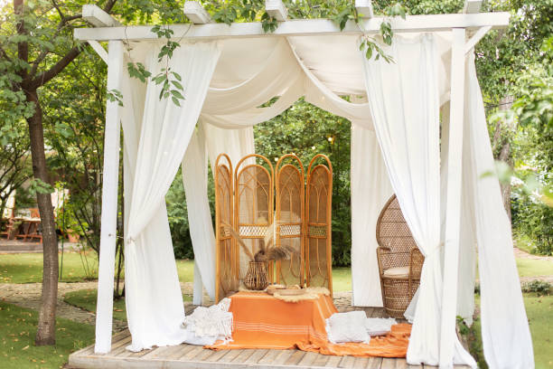istock Summer gazebo with flowing white curtains. Wedding boho decoration. Decor outdoor terrace with wicker furniture. Outdoor design of arbour. Rattan peacock armchair and decorative folding screen divider 1640312449